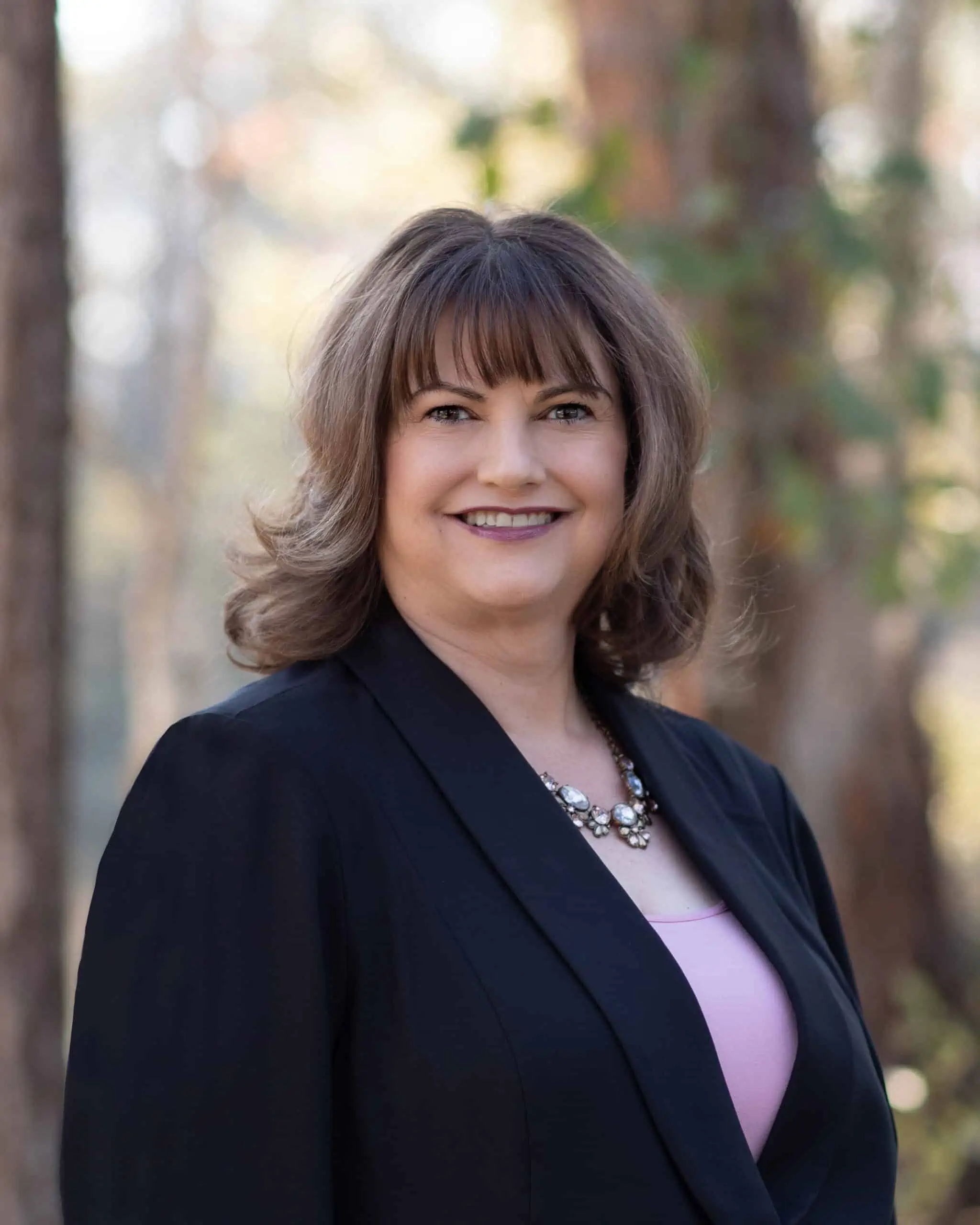 A woman in a black blazer and pink top smiling in the woods.