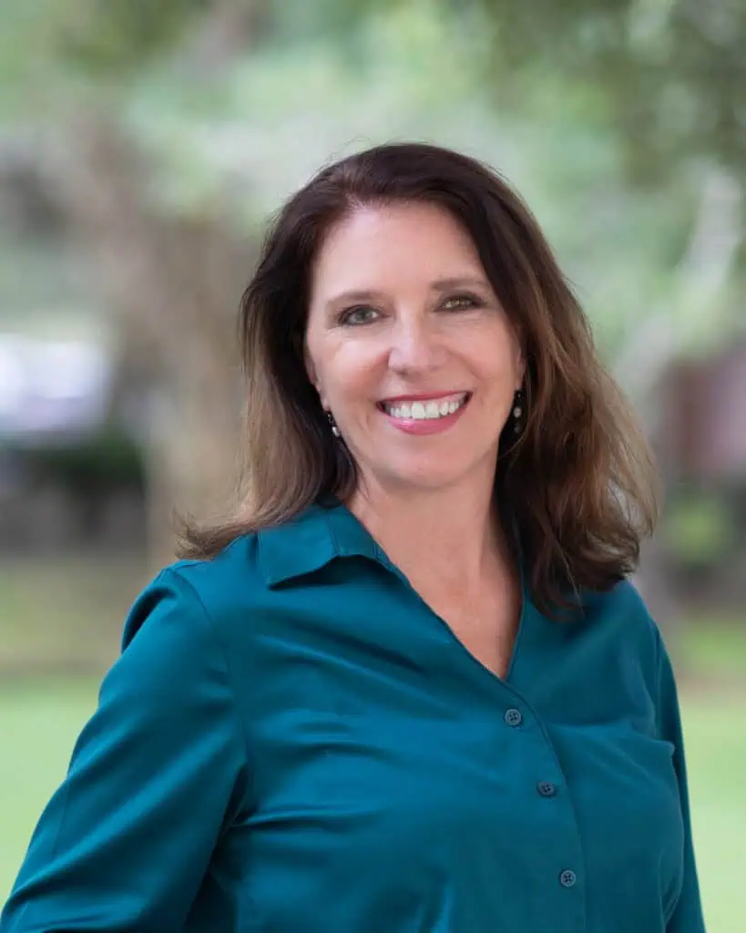 A woman with long brown hair, wearing a teal blouse, smiles outdoors with blurred greenery in the background.