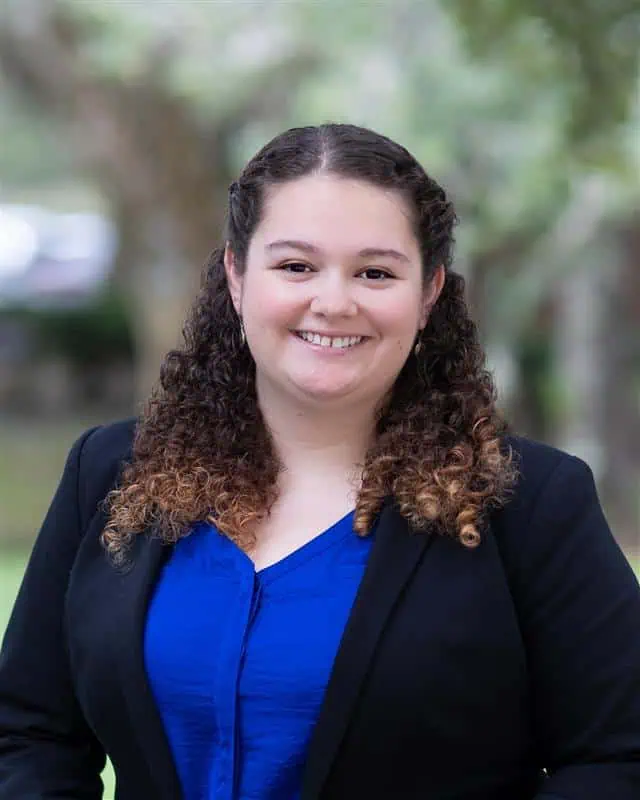 A person with curly hair wearing a blue shirt and black blazer stands outdoors, smiling at the camera.