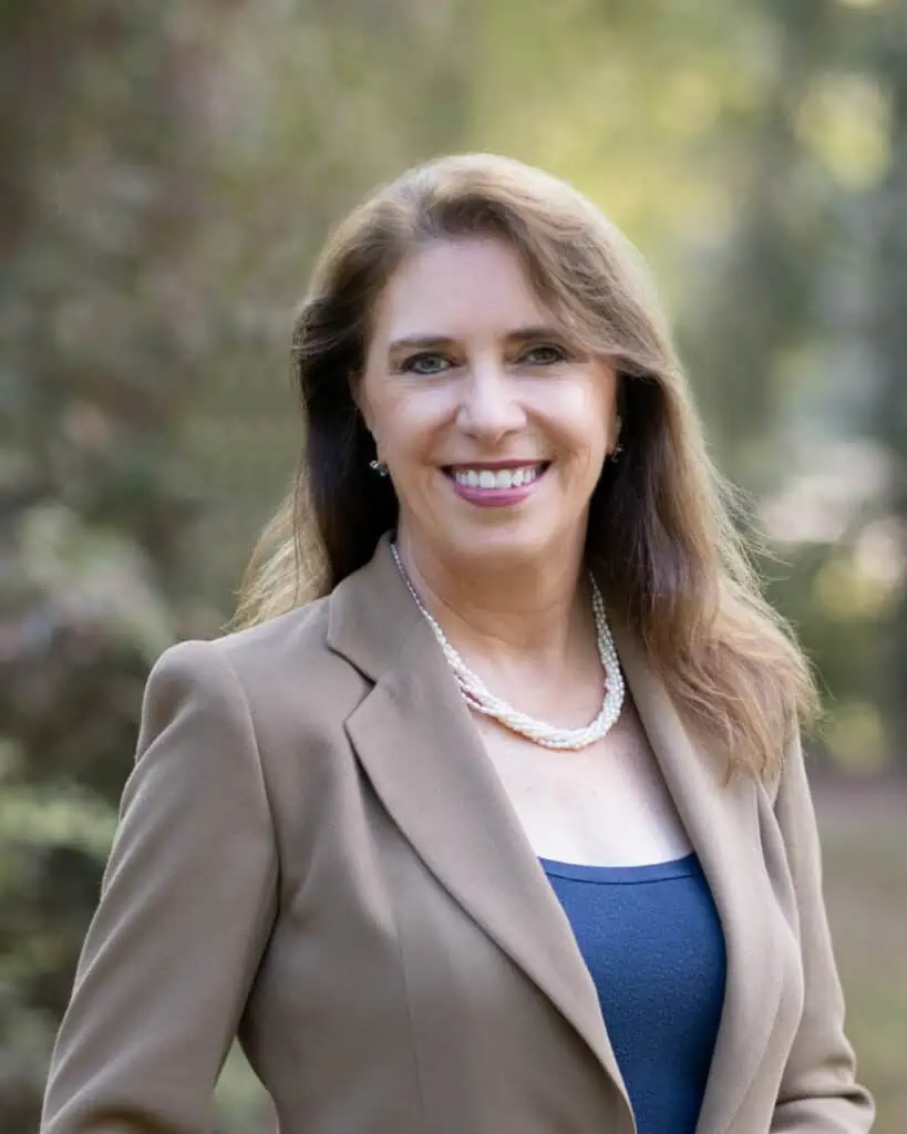 A woman in a tan jacket smiling in the woods.