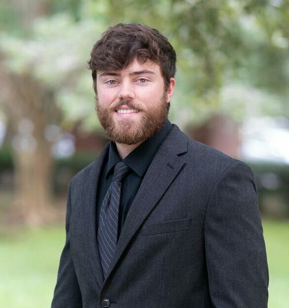 A man with a beard wearing a black suit and tie stands outdoors with trees in the background.