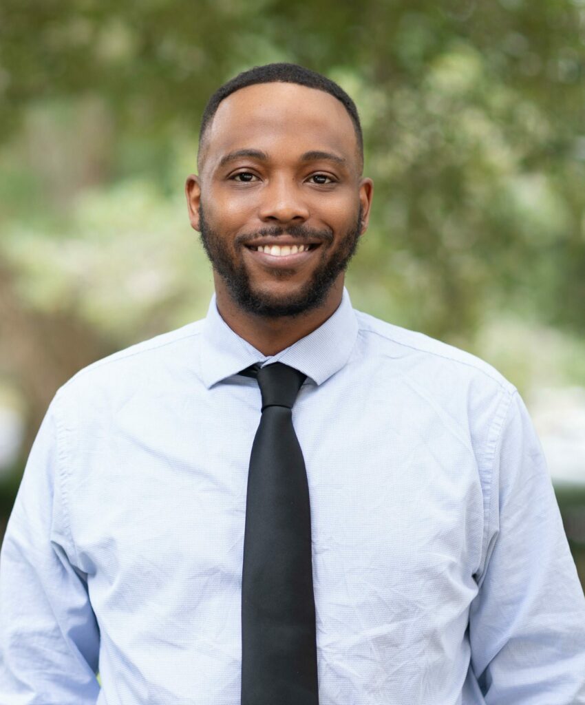 A person smiling, wearing a light blue shirt and black tie, standing outdoors with blurred greenery in the background.