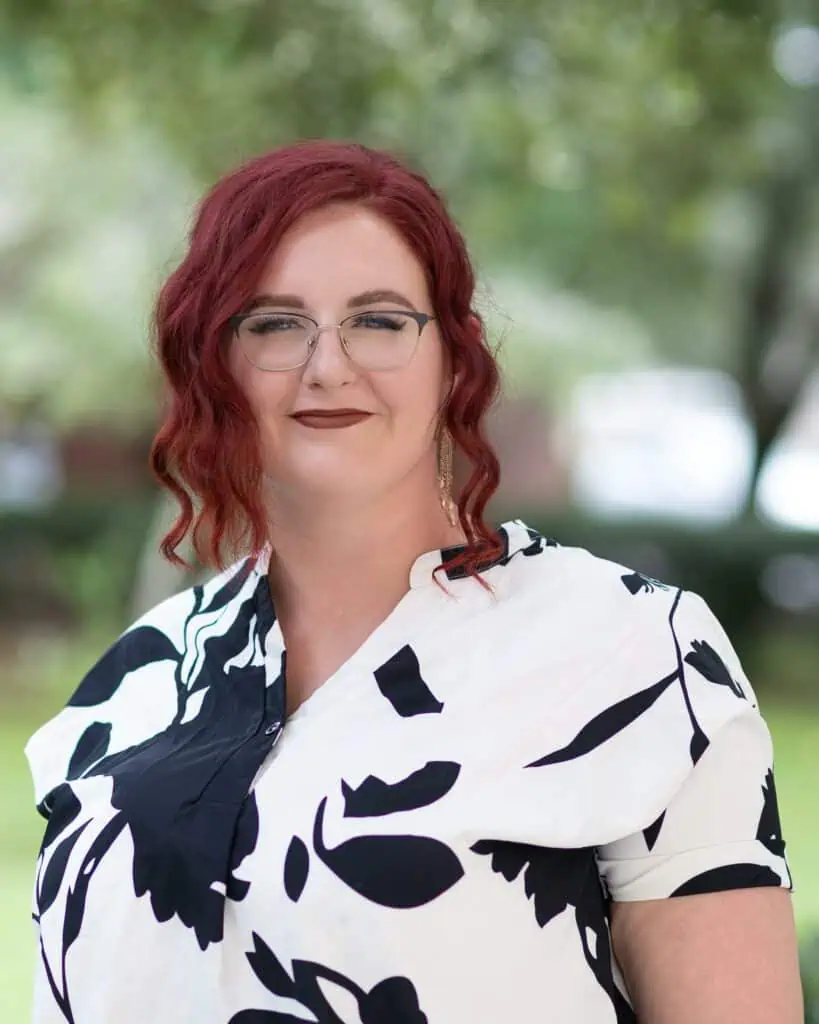 A person with red hair and glasses is wearing a black and white patterned shirt, standing outdoors with trees blurred in the background.