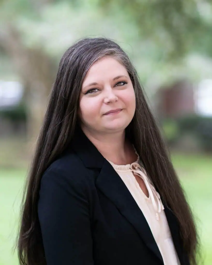 Woman with long brown hair wearing a black blazer over a light blouse, standing outdoors with a blurred green background.