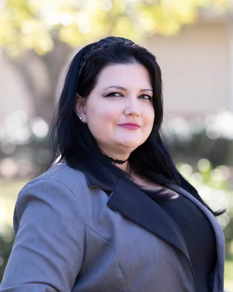 Woman with long dark hair wearing a gray blazer and black top, standing outdoors with a blurred background.