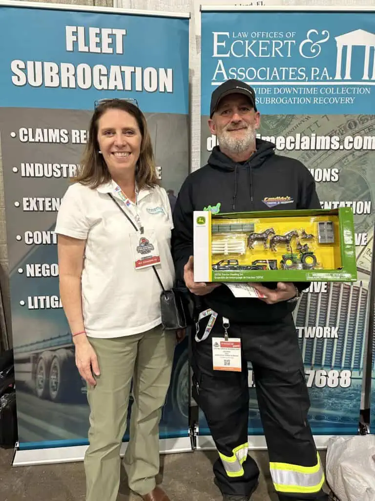 Two individuals at an expo stand, one holding a model truck, with a banner displaying corporate services in the background.