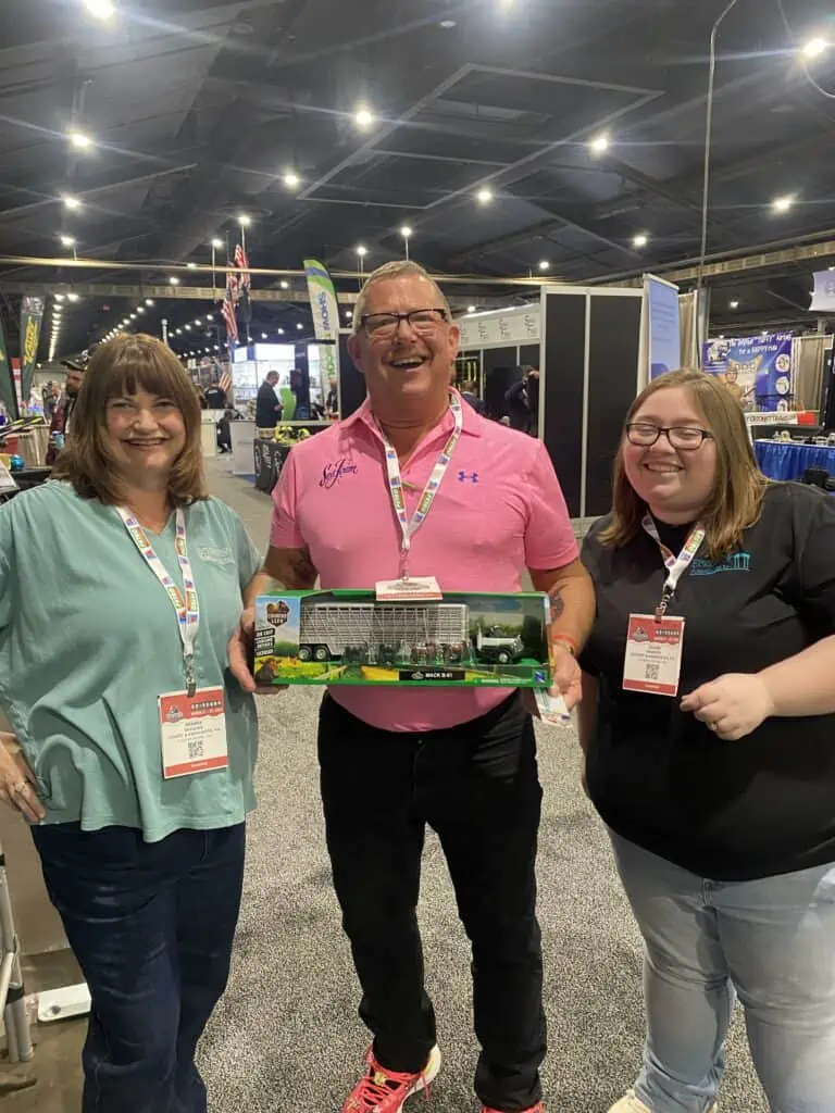 Three individuals smiling at a convention, holding a model train.