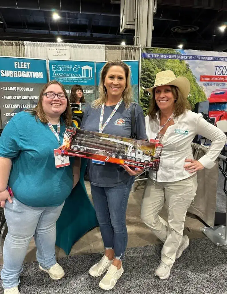 Three individuals at a trade show or event, one holding a model train, smiling for the camera.