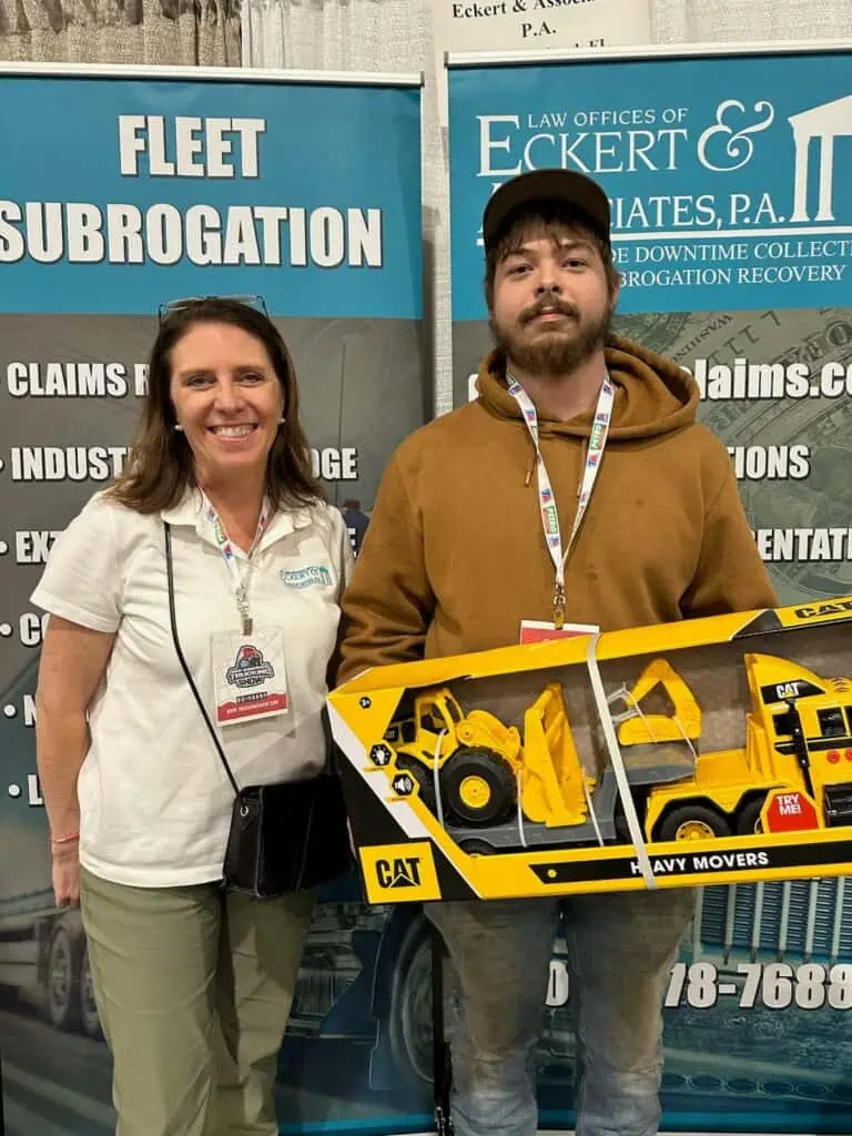 Two individuals posing for a photo at an event booth, one holding a model construction vehicle toy.