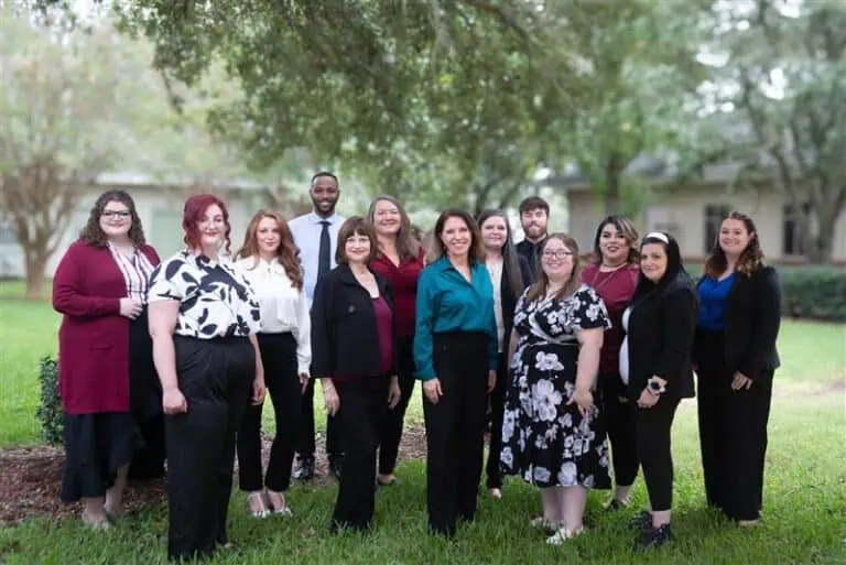 A group of people stands together outside in a park-like setting, with trees in the background. They are dressed in business casual attire.