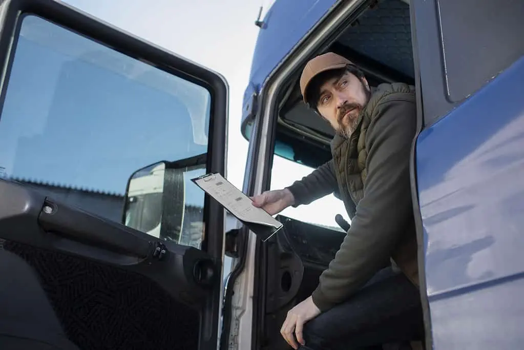 A man sits in the cab of a truck with the door open, holding a clipboard and looking outside.