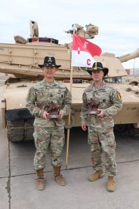 Two soldiers in camouflage uniforms and black hats stand in front of a military tank, holding awards, with a red and white flag marked "C 5" between them.