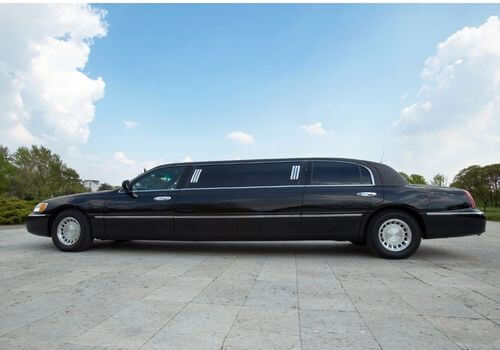 A black stretch limousine is parked on a paved surface with trees and blue sky in the background.