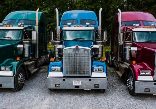 Three semi-trailer trucks—green, blue, and red—are parked side by side on a gravel lot with trees in the background.