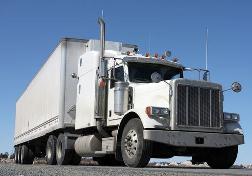 A white semi-truck with a large trailer is parked on a gravel surface under a clear blue sky.