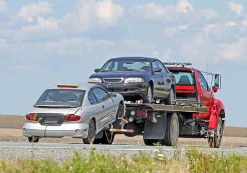 A red tow truck is transporting a damaged silver car on its flatbed and towing another silver car behind it on a highway.