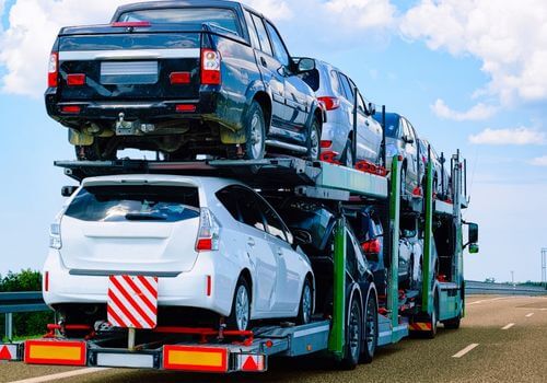 A car carrier truck transports multiple vehicles stacked in two rows on a highway under a blue sky with clouds.