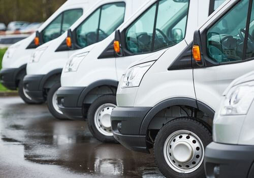 A row of white delivery vans parked side by side on a wet pavement, showing the front and side views of the vehicles.