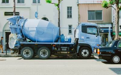 A blue cement mixer truck is parked on a city street beside a black car, with buildings and trees in the background.