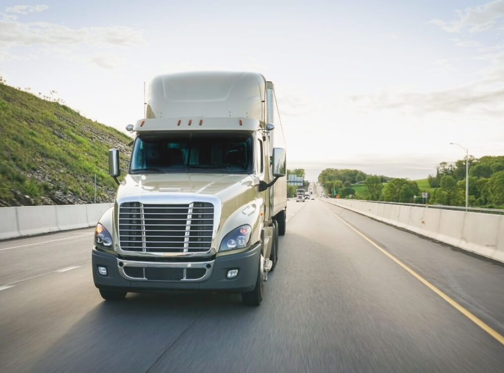 A beige semi-truck drives on a divided highway bordered by a grassy slope and trees under a partly cloudy sky.