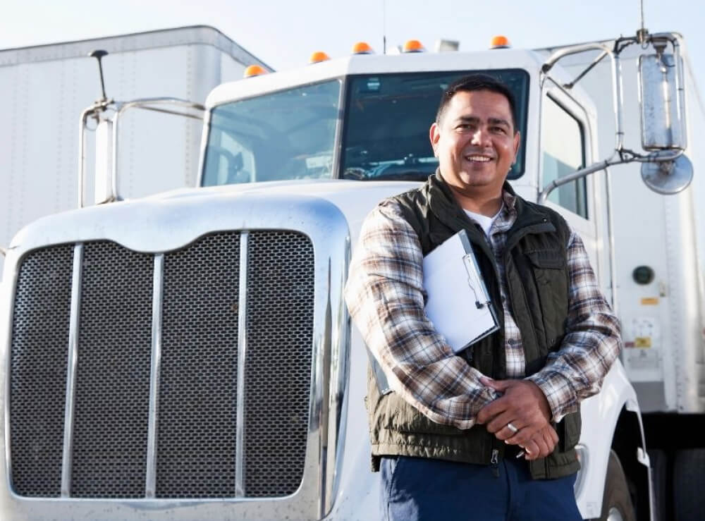 A man holding a clipboard stands in front of a white semi-truck, smiling at the camera.