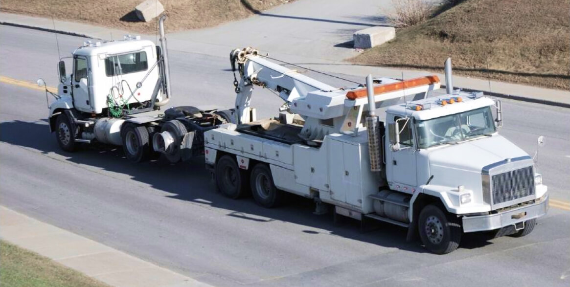 A large tow truck is towing a white semi-truck cab along a paved road on a sunny day.