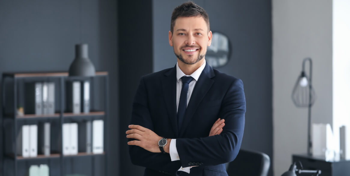A man in a navy suit and tie stands with arms crossed, smiling in a modern office with shelves, folders, and decor in the background.