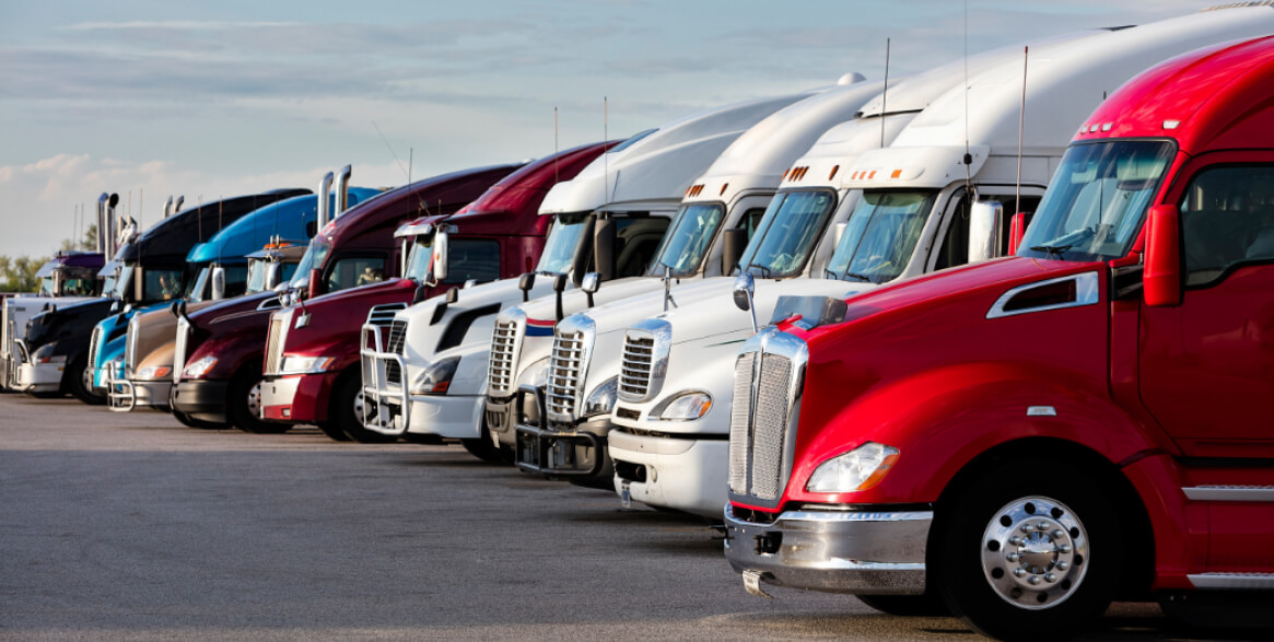 A row of parked semi-trucks in various colors is lined up in a lot under a partly cloudy sky.