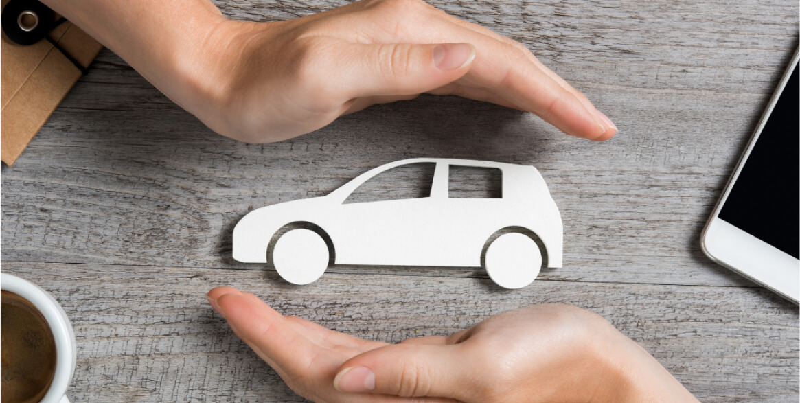 Two hands form a protective gesture around a white car cutout on a wooden table, next to a cup of coffee and a smartphone.