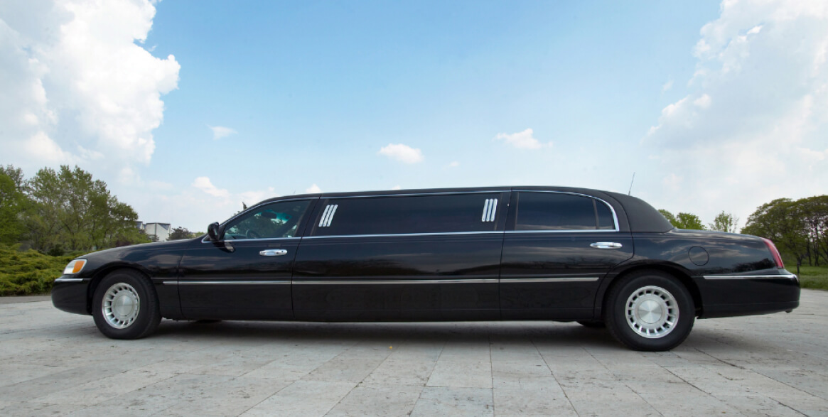 A long black stretch limousine is parked on a paved surface outdoors, with trees and a partly cloudy sky in the background.