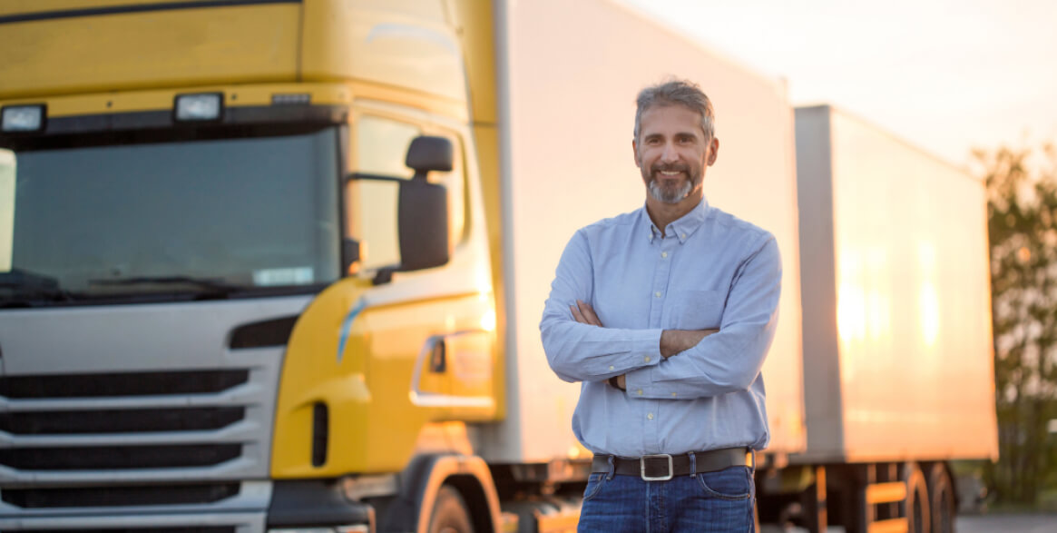 Middle-aged man standing with arms crossed in front of a yellow and white semi-truck, outdoors in daylight.