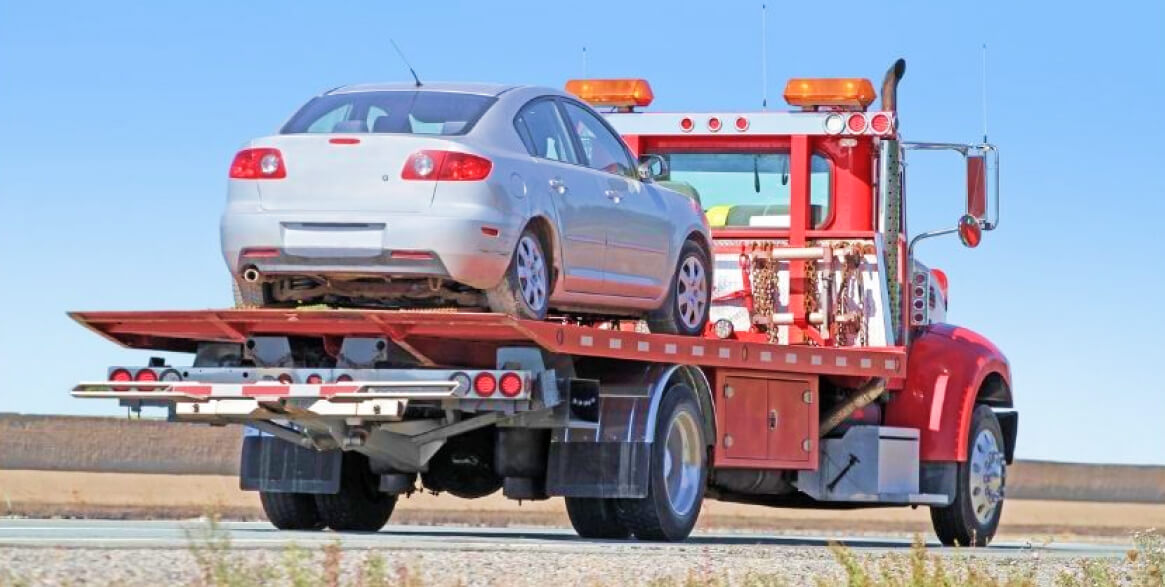 A silver sedan is loaded onto the flatbed of a red tow truck on a clear day with an open landscape in the background.