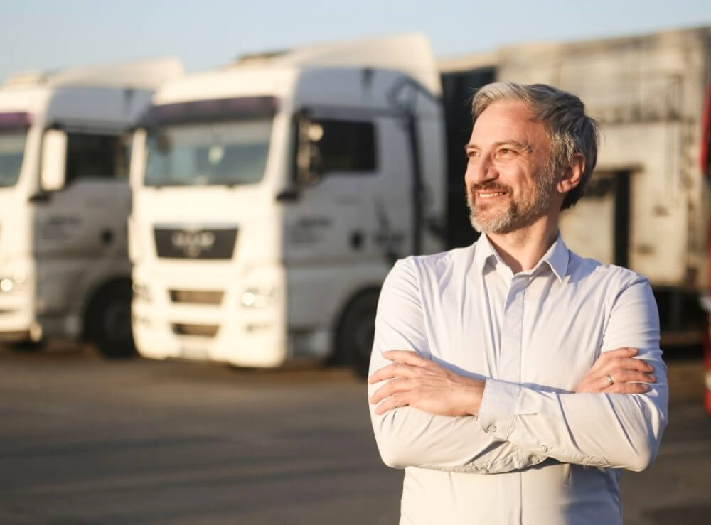 A man in a white shirt stands with arms crossed, smiling, in front of several parked white semi-trucks in a lot.