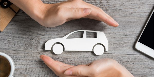 Two hands form a protective gesture around a white car cutout on a wooden table, next to a cup of coffee and a smartphone.